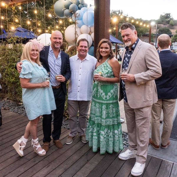 Five smiling guests holding drinks, posing outdoors on a wooden deck with string lights and balloons.