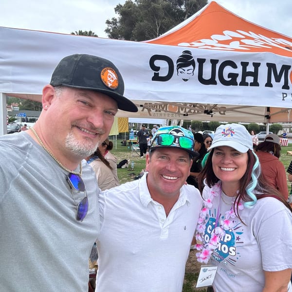 Three smiling people pose in front of a Dough Momma Pizzeria tent at an outdoor event.