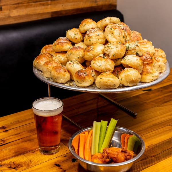 A tiered tray of Dough Momma Pizzeria garlic knots, a beer, and a bowl of wings with carrots and celery.