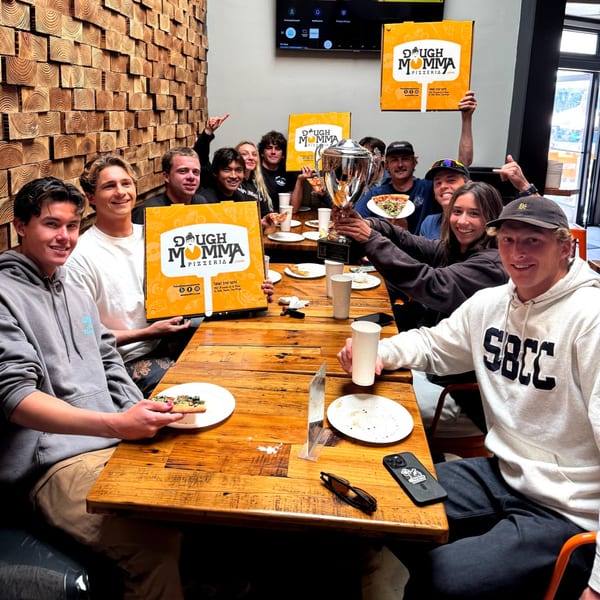 A group of young people celebrating with pizza, Dough Momma boxes, and a trophy at a wooden table.