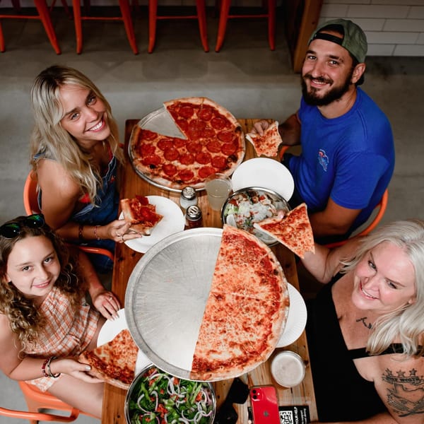 Happy family enjoying Dough Momma Pizzeria's pepperoni and cheese pizzas, with salads and drinks.