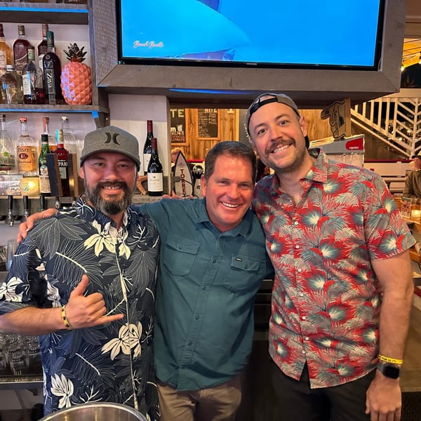 Three smiling men in casual shirts pose together in front of a bar with bottles and a TV.