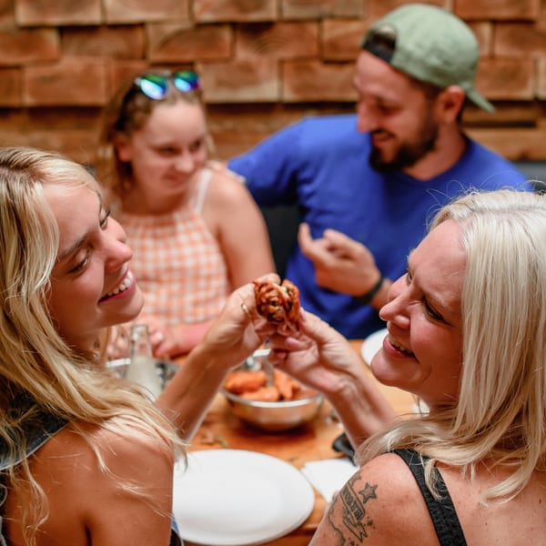 Two women smiling and sharing a chicken wing at a table with friends at Dough Momma Pizzeria.
