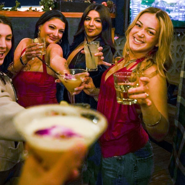 Four smiling women holding cocktails and drinks, enjoying a night out at Dough Momma Pizzeria.