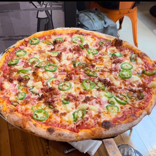 A large Dough Momma Pizzeria pizza with pineapple, ham, bacon, and jalapeños on a wooden table.