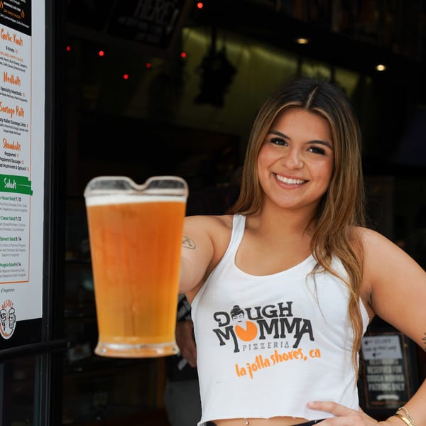 A smiling woman in a Dough Momma Pizzeria shirt holds out a glass of beer.