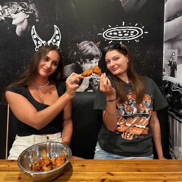 Two women holding up chicken wings from a metal bowl, smiling in a pizzeria with quirky wall art.